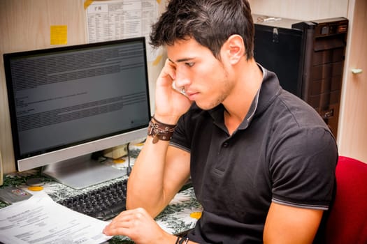 Waist Up Closeup of Young Attractive Man with Dark Hair Sitting at Computer Desk Talking on Cell Phone in Dorm Room