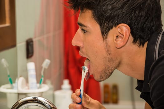 Headshot of attractive young man brushing teeth and cleaning tongue with toothbrush, looking at himself in mirror