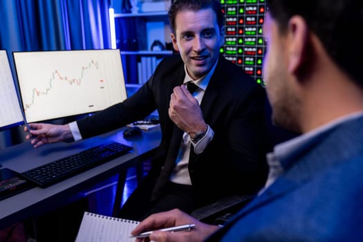 Two stock exchange traders discussing on dynamic investment graph, trending of high profit in currency stock. Showing financial benefit data on monitor screen in neon light at workplace. Sellable.