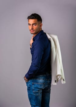 A man in a blue shirt and jeans with a jacket on his back. Photo of a man wearing casual clothing with a jacket slung over his shoulder in studio shot on grey background