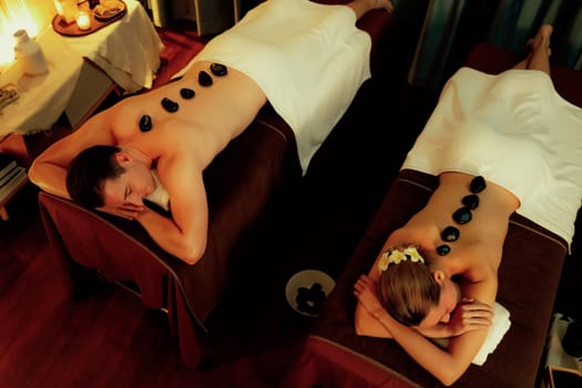 Hot stone massage at spa salon in luxury resort with warm candle light, blissful couple customer enjoying spa basalt stone massage glide over body with soothing warmth. Quiescent