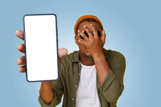Studio shot of Exited happy young african handsome with sign of facepalm shows mobile phone screen with empty copy space on white display for your text. Man in basic closes isolated on blue background. High quality photo