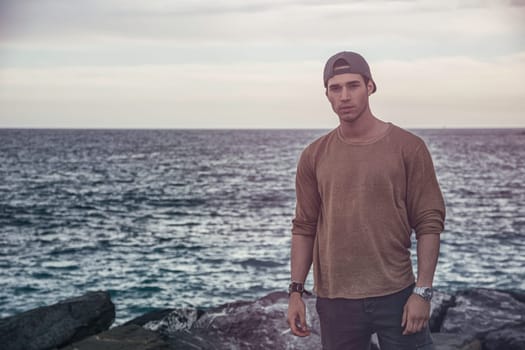 A young attractive man standing on top of a rocky beach next to the ocean or sea in autumn or winter