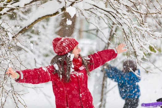 Happy girl in red jumpsuit, knocking down branches with snow and laughing.