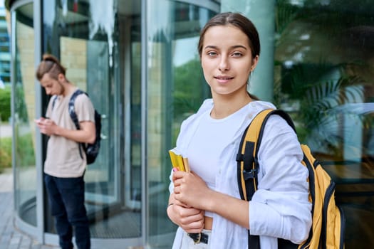 Portrait of high school student, smiling girl with backpack looking at camera, outdoor, educational building background. Adolescence, 15, 16, 17 years old, lifestyle, education concept