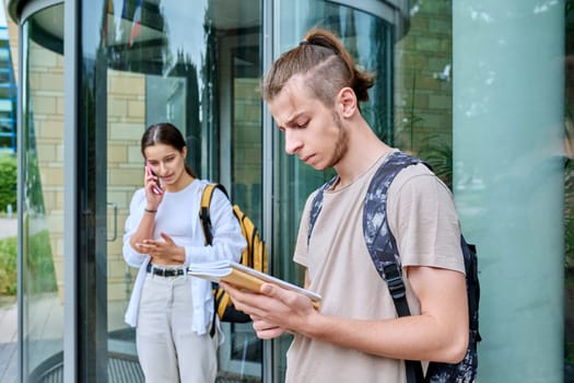 Hipster guy teenager student 18, 19 years old with backpack notebooks standing outdoor, educational building background. Youth, education, lifestyle concept