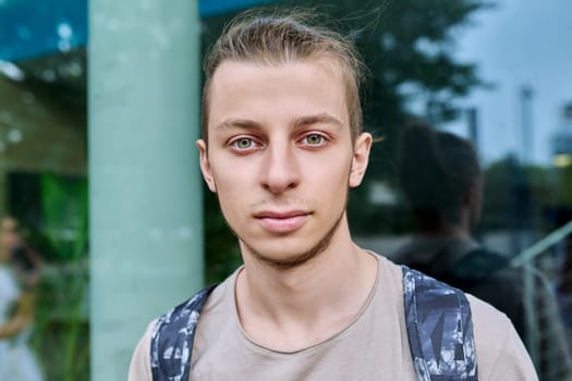 Headshot portrait of confident college student guy with backpack, looking at camera, outdoor, educational building background. Youth, education, lifestyle concept