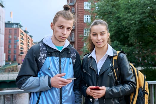 Outdoor portrait of teenagers students friends smiling guy and girl with smartphones looking at camera on street of modern city. College, 17-19 years old, urban lifestyle, adolescence youth concept