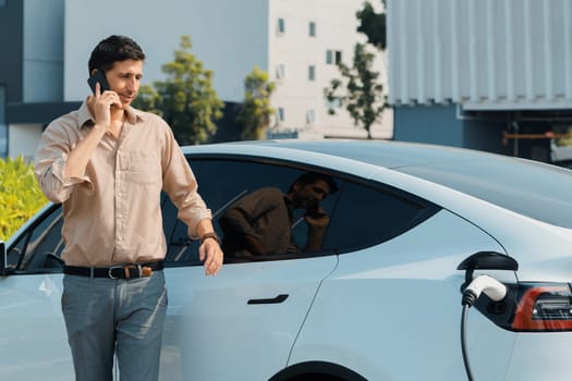 Young man recharge EV electric vehicle at green city commercial mall parking lot while talking on phone. Sustainable urban lifestyle for eco friendly EV car with battery charging station. Expedient