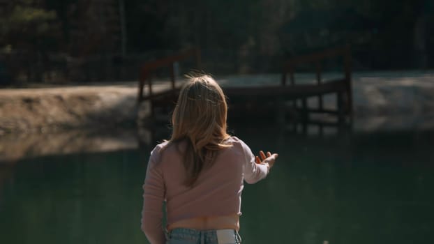 Rear view of young woman throwing rocks into water. Stock footage. Attractive young woman sits on pier and throws stones. Woman throws stones on shore of lake on sunny summer day.