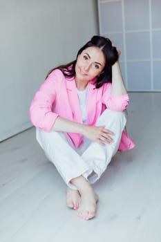 Beautiful fashionable brunette woman sitting on the floor in pink jacket and white pants