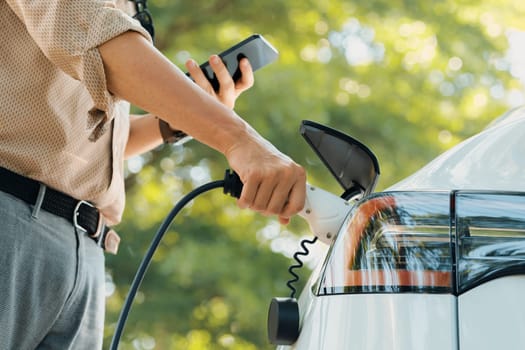 Young man use smartphone to pay for electricity at public EV car charging station green city park. Modern environmental and sustainable urban lifestyle with EV vehicle. Expedient