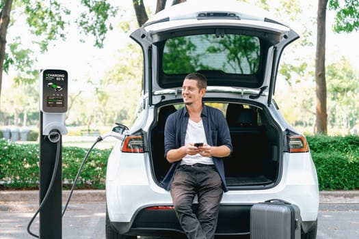 Man using smartphone online banking application to pay for electric car battery charging from EV charging station during vacation holiday road trip at national park or summer forest. Exalt