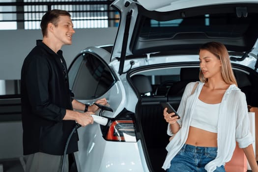 Young couple travel with EV electric car to shopping center parking lot charging in downtown city showing urban sustainability lifestyle by green clean rechargeable energy of electric vehicle innards