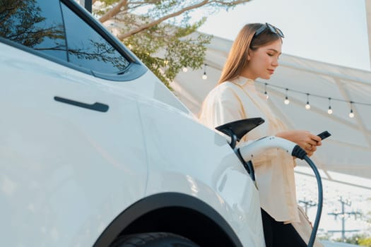 Sustainable urban commute with young woman pay electricity for EV electric car recharging at outdoor cafe in springtime garden, green city sustainability and environmental friendly EV car. Expedient