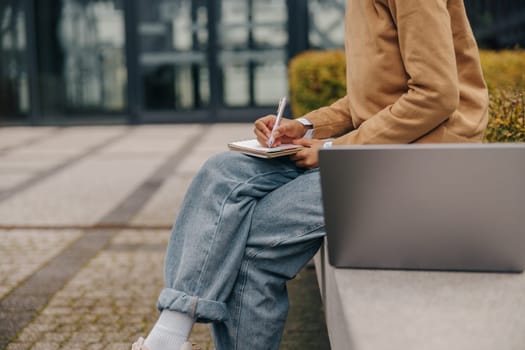 Female freelancer working laptop and making notes sitting outside on background of office building