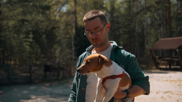 Man holds dog in hands in woods. Stock footage. Man holds Jack Russell terrier in arms. Man holds dog in arms in park on sunny summer day.