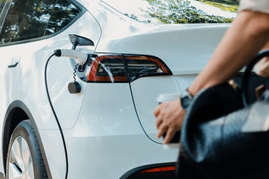 Young man recharge electric car's battery from charging station in outdoor green city park in springtime. Rechargeable EV car for sustainable environmental friendly urban travel lifestyle. Expedient