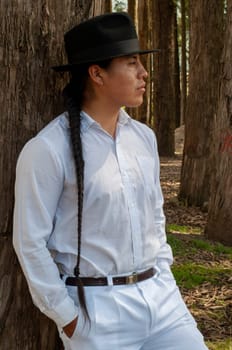 vertical photo of a south american boy leaning against a tree wearing a white suit, black hat and long braid. High quality photo