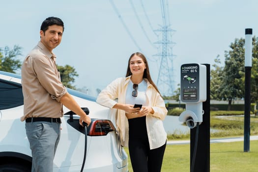 Young couple use smartphone to pay for electricity at public EV car charging station green city park. Modern environmental and sustainable urban lifestyle with EV vehicle. Expedient