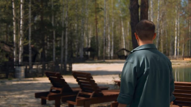 Man walks by lake with places to relax. Stock footage. Man walks along forest of resort lake in forest in summer. Beautiful lake with recreation center on sunny summer day.
