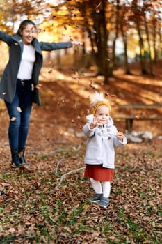 Laughing mother throws up fallen leaves on a little girl in the autumn forest. High quality photo