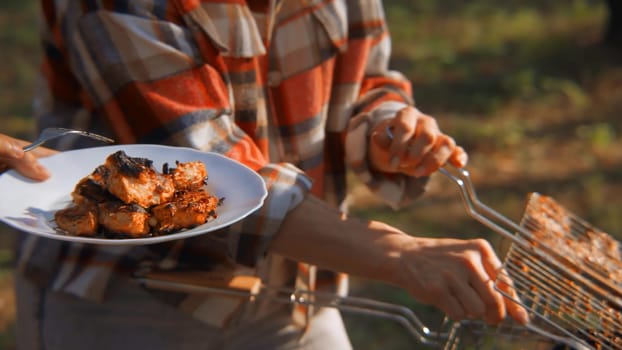 Close-up of grilled chicken pieces put on plate. Stock footage. Grilled chicken is put in plate from barbecue in nature. Ready-made barbecue chicken in forest on sunny summer day.