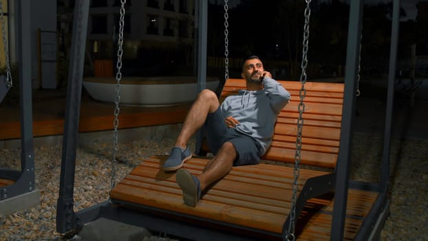 Young man on swing talking on phone. Media. Happy young man is answering the telephone on swing. Young man is sitting in modern park and answering telephone with beloved.