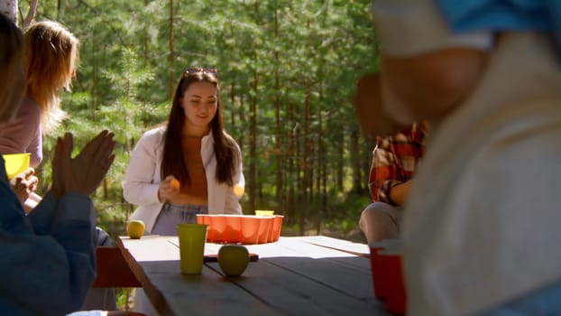 Young people play beer pong. Stock footage. Game for group of friends with glasses of alcohol and ping pong ball. Friends play beer pong in nature in summer.