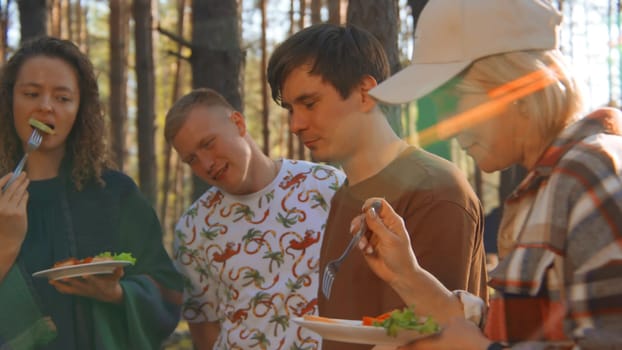 Group of friends eats vegetables at grill in nature. Stock footage. Beautiful friends have fun chatting at barbecue in forest. Fun vacation with friends in forest on sunny summer day.
