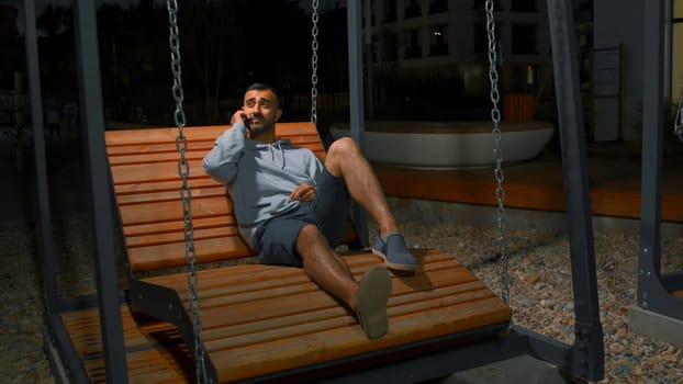 Young man on swing talking on phone. Media. Happy young man is answering the telephone on swing. Young man is sitting in modern park and answering telephone with beloved.