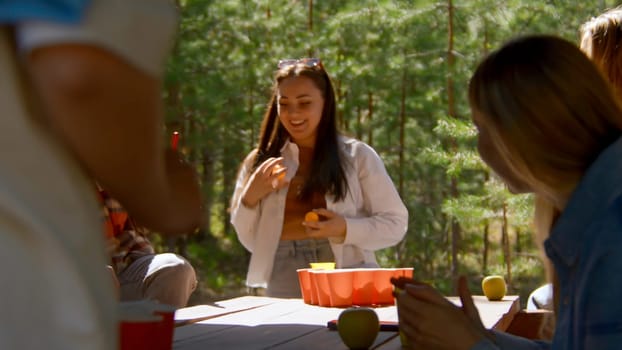 Young people play beer pong. Stock footage. Game for group of friends with glasses of alcohol and ping pong ball. Friends play beer pong in nature in summer.