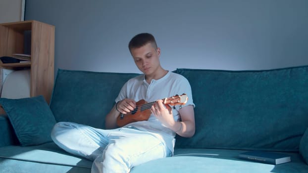 Young man plays ukulele at home. Media. Man plays ukulele alone at home. Young man practices playing small guitar while sitting on couch.