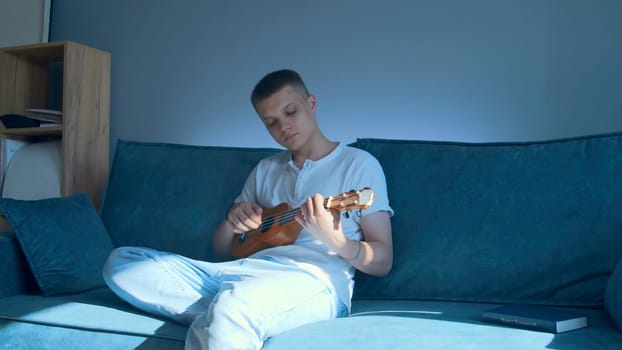 Young man plays ukulele at home. Media. Man plays ukulele alone at home. Young man practices playing small guitar while sitting on couch.