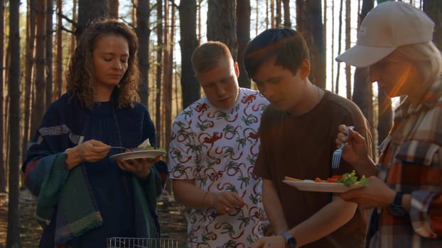 Friends eat barbecue from grill in woods. Stock footage. Friends relax in nature at barbecue and eat. Friends eat cooked meat on grill on sunny summer day.