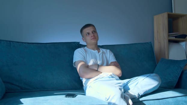 Young man is sitting on sofa and waiting for phone call. Media. Handsome young man is waiting for call sitting next to phone. Man is sitting on couch next to phone at home.