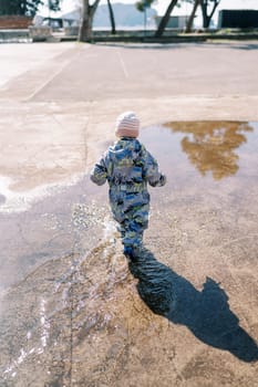 Little girl in overalls walks through a puddle, making splashes. Back view. High quality photo
