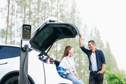 Lovely young couple recharging battery for electric car while relaxing and sitting on the trunk during road trip travel EV car in autumnal forest. Eco friendly travel on vacation during autumn. Exalt