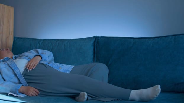 Young woman lies down on sofa with tired look. Media. Young woman lies down on sofa after hard day. Woman in tired clothes lies down on sofa at home.