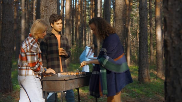 Friends cook grilled meat in forest in summer. Stock footage. Beautiful friends are relaxing in nature with barbecue in forest. Relaxing with friends in nature with barbecue on sunny summer day.