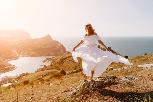 Happy woman in a white dress and hat stands on a rocky cliff above the sea, with the beautiful silhouette of hills in thick fog in the background