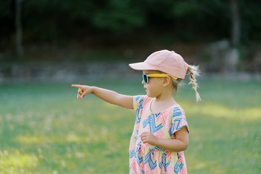 Little girl in sunglasses stands in the park and points into the distance. Side view. High quality photo