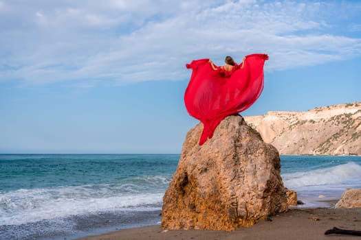 woman sea red dress. Woman with long hair on a sunny seashore in a red flowing dress, back view, silk fabric waving in the wind. Against the backdrop of the blue sky and mountains on the seashore