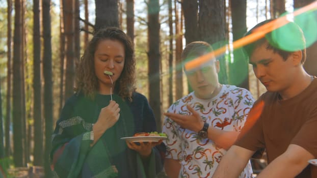 Friends communicate and eat food from barbecue in nature in summer. Stock footage. Friends are having fun talking and eating in woods on sunny summer day. Bright cheerful communication of friends in nature with barbecue.
