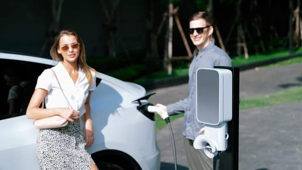 Young couple travel with EV electric car charging in green sustainable city outdoor garden in summer shows urban sustainability lifestyle by green clean rechargeable energy of electric vehicle innards