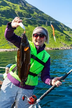 A smiling woman is seen fishing in the Norwegian Sea, holding a large catch. The tranquil scene showcases the enjoyable pastime of fishing in the serene surroundings of the sea.