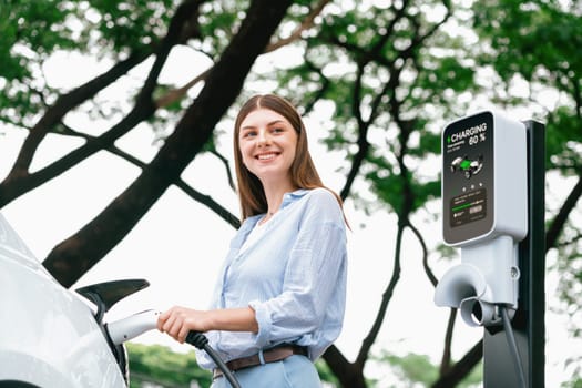 Young woman recharging battery for electric car during road trip travel EV car in natural forest or national park. Eco friendly travel during vacation and holiday. Exalt