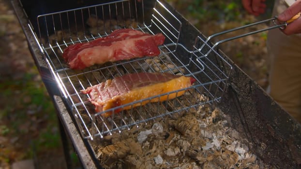 Close-up of man cooking juicy pieces of meat on grill. Stock footage. Man cooks two delicious pieces of red meat on coals. Grilling meat outdoors in summer.