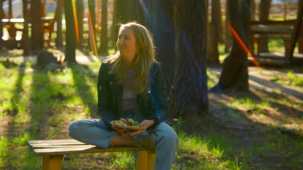 Young woman is sitting on bench and eating barbecue. Stock footage. Beautiful woman is sitting alone and eating barbecue in park in summer. Woman with barbecue plate in nature on sunny summer day.