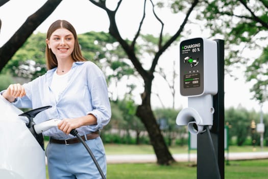 Young woman recharge EV electric vehicle's battery from EV charging station in outdoor green city park scenic. Eco friendly urban transport and commute with eco friendly EV car travel. Exalt